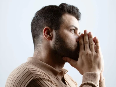 Hombre de perfil con barba y suéter beige cubriéndose la boca y nariz con las manos, posiblemente por un estornudo o tos debido a una alergia.