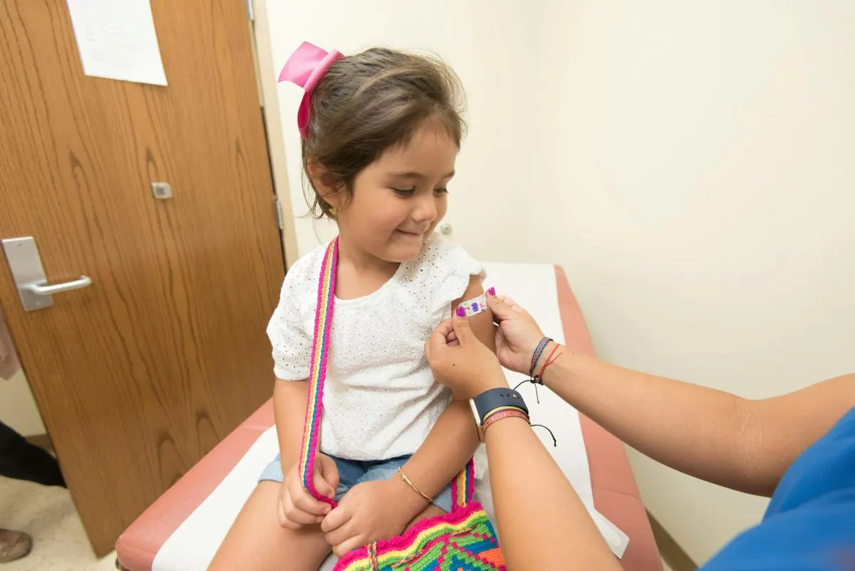 Niña pequeña sonriendo mientras una enfermera le coloca una curita en el brazo, representando la importancia de las vacunas para entrenar al sistema inmunológico desde la infancia.