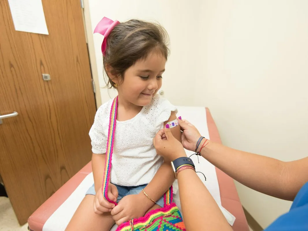 Niña pequeña sonriendo mientras una enfermera le coloca una curita en el brazo, representando la importancia de las vacunas para entrenar al sistema inmunológico desde la infancia.